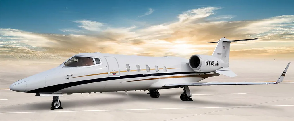 A white Learjet 75 business jet with black and gold stripes is parked on a tarmac under a dramatic sky.