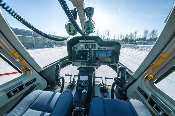 The cockpit of a helicopter shows the instrument panel with multiple screens, controls, and blue seats, looking out onto a snowy field.