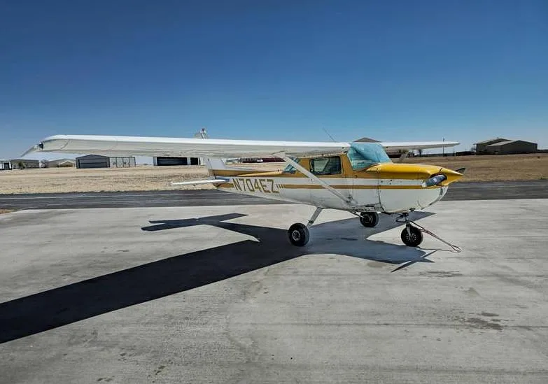 A white and yellow Cessna 150, registration N704EZ, parked on a tarmac with a windshield sunshade.