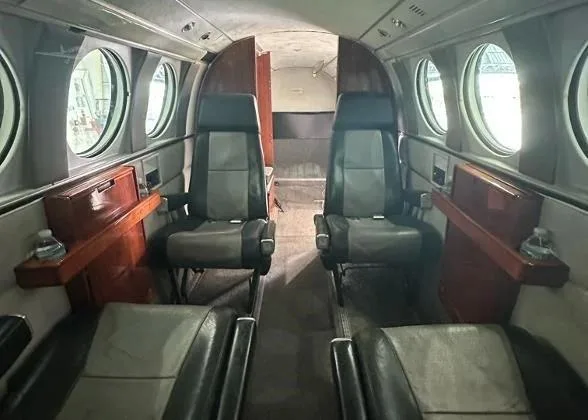 The aircraft's cabin interior shows grey and black leather seats, wooden side tables, and round windows, viewed from the rear.