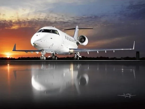 A white business jet, likely a Bombardier Challenger, is parked on a reflective surface at sunset.