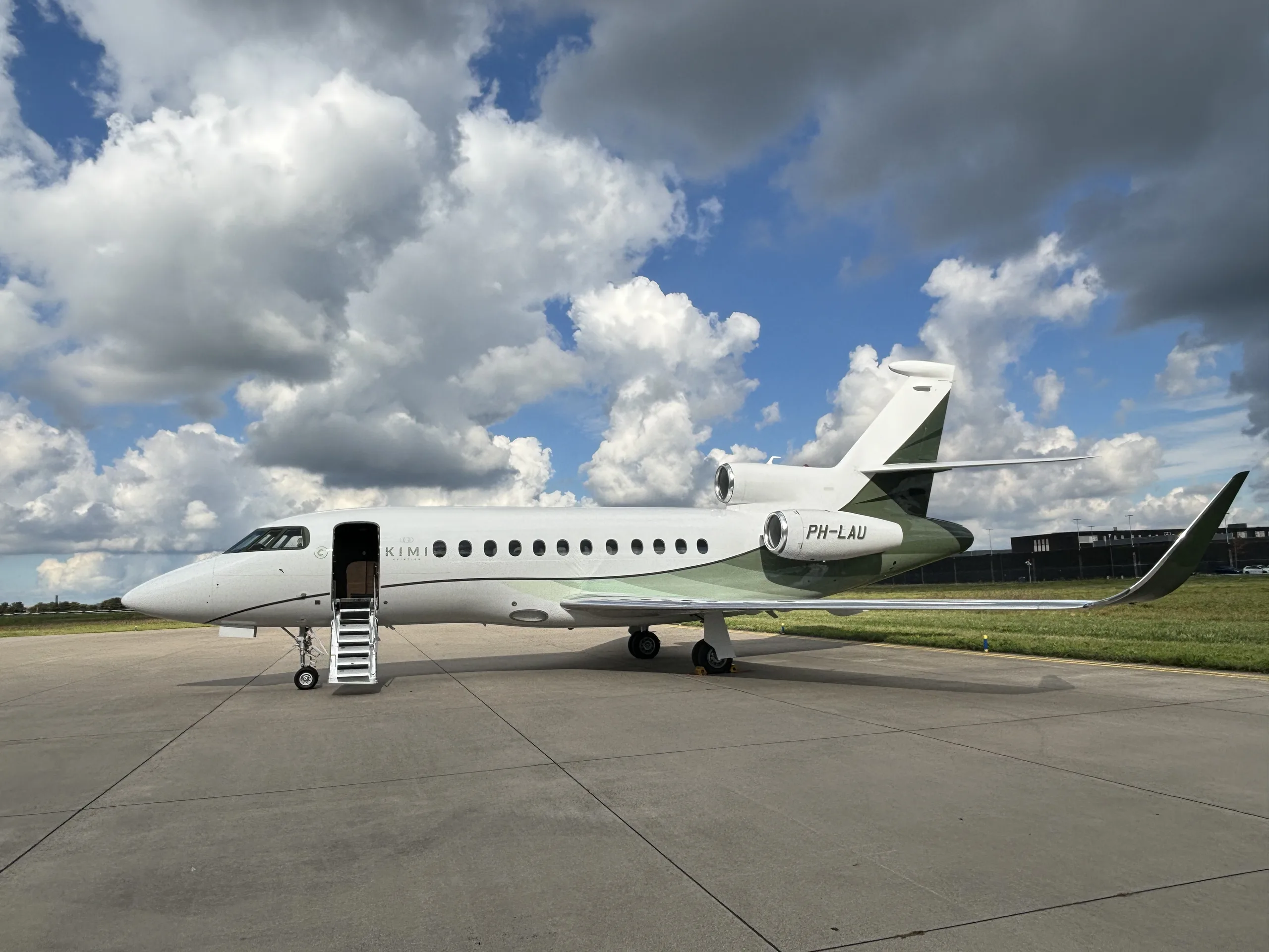 A white business jet with green accents on the tail and engines, registration PH-LAU, parked on an airfield under a cloudy sky.