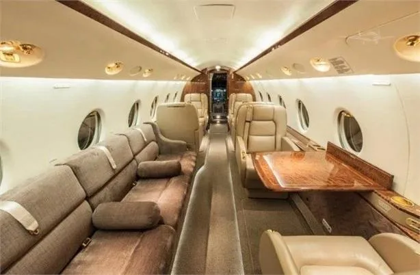A wide view of a private jet's luxurious cabin interior with a long beige sofa on the left and two beige leather seats with a wood-grain table on the right.