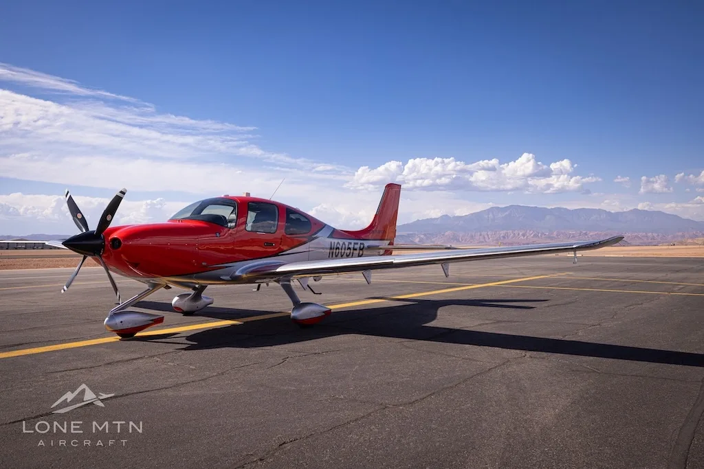 A red and silver Cirrus SR22T aircraft (N605FB) parked on an airfield under a blue sky with mountains in the background.