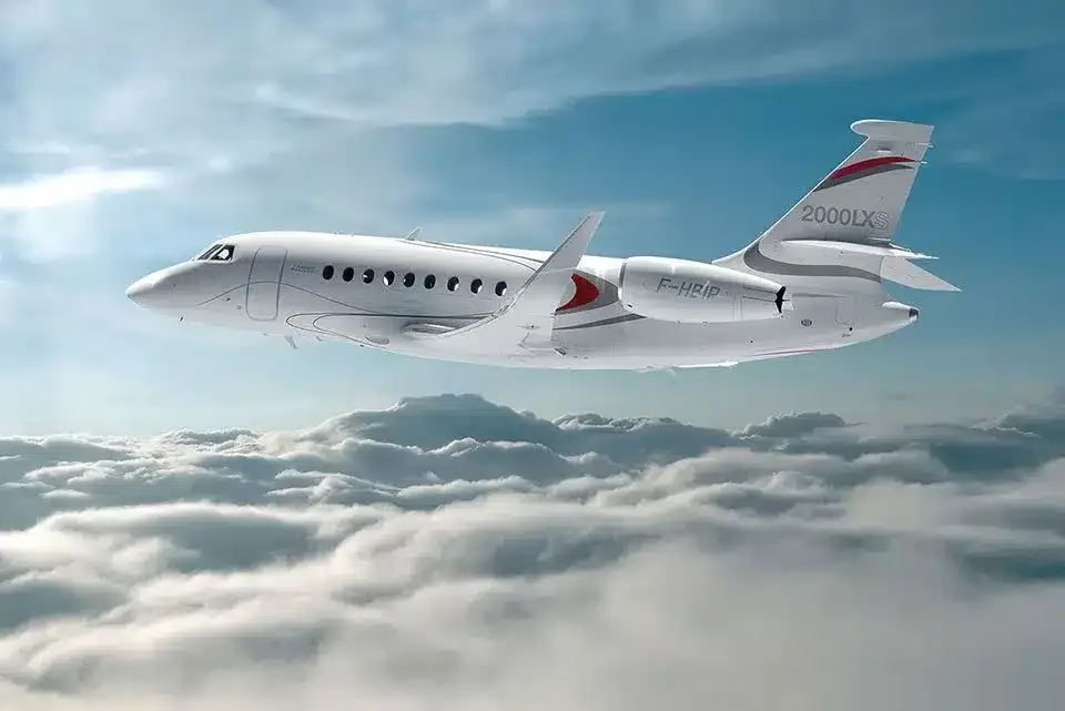 A white Dassault Falcon 2000LXS business jet with red and grey tail accents flies above a layer of clouds.