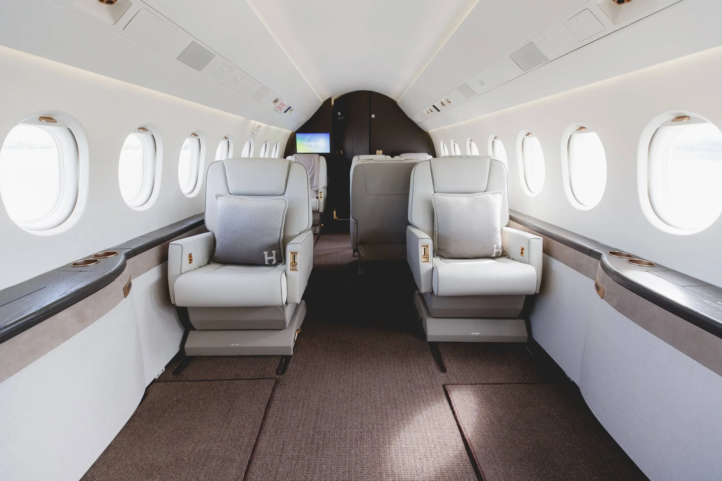 The luxurious interior cabin of a private jet, featuring two rows of light-colored leather seats with "H" monogrammed pillows.