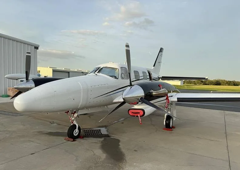 A white and black twin-engine propeller aircraft, possibly a Piper Navajo, parked on a tarmac with hangars in the background.