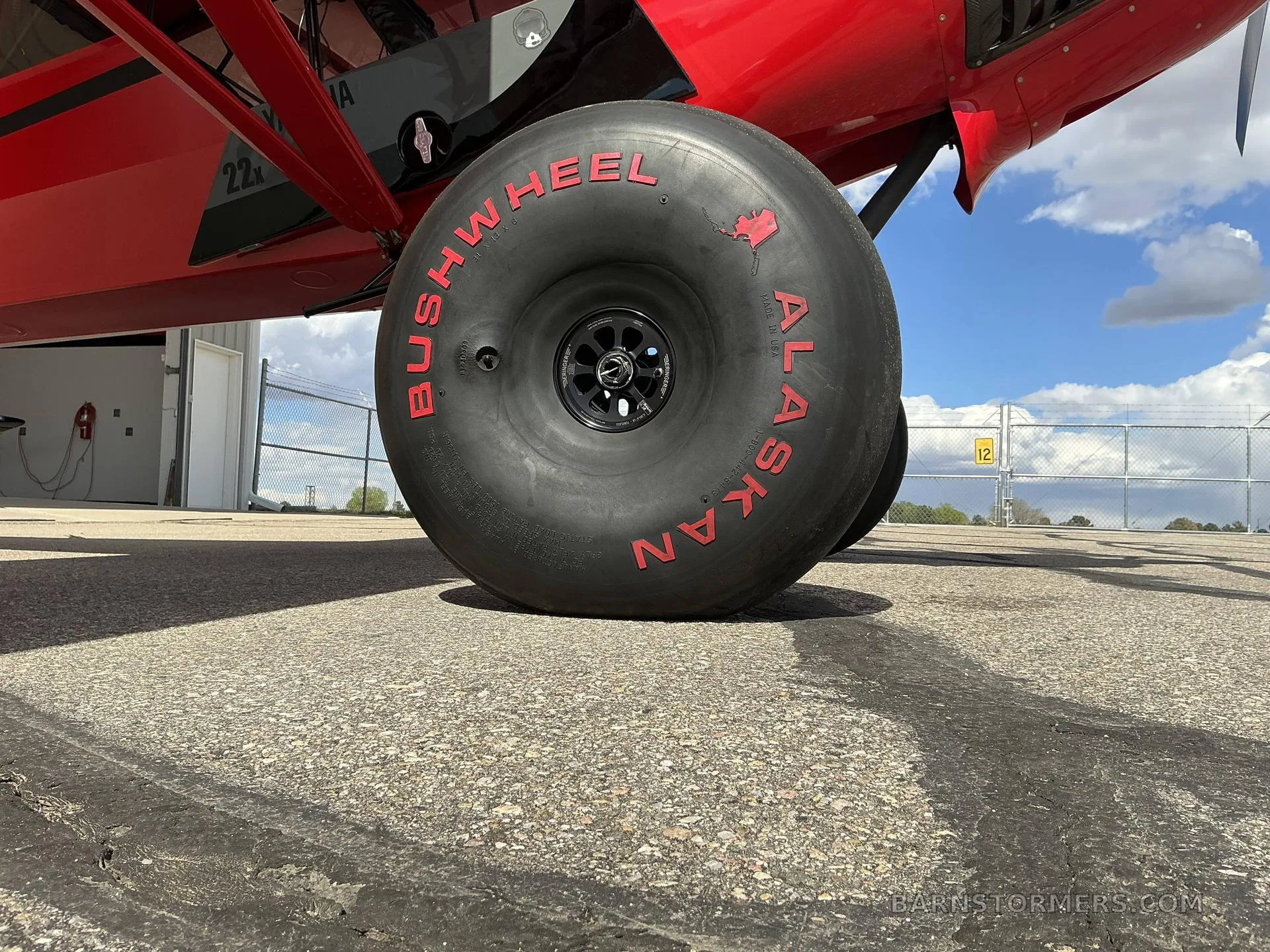 A close-up of a large black "Bushwheel Alaskan" tire with red lettering on a red aircraft, parked on an asphalt surface.