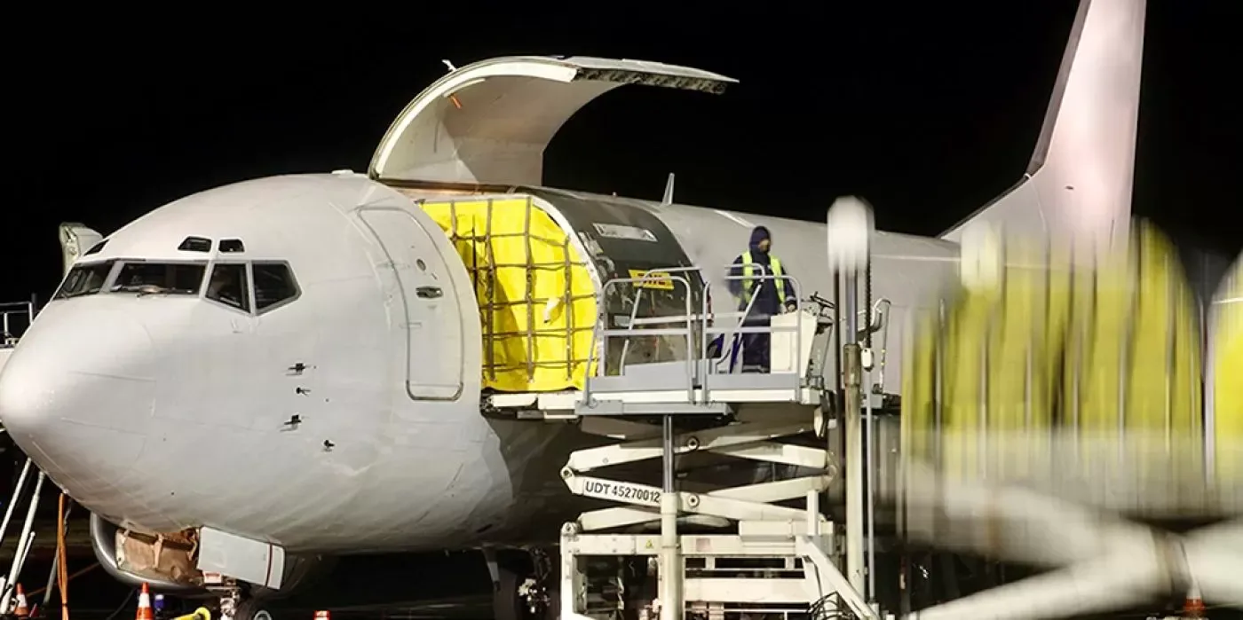 The front section of a white cargo plane with its nose door open, being loaded by a worker on a lift at night.