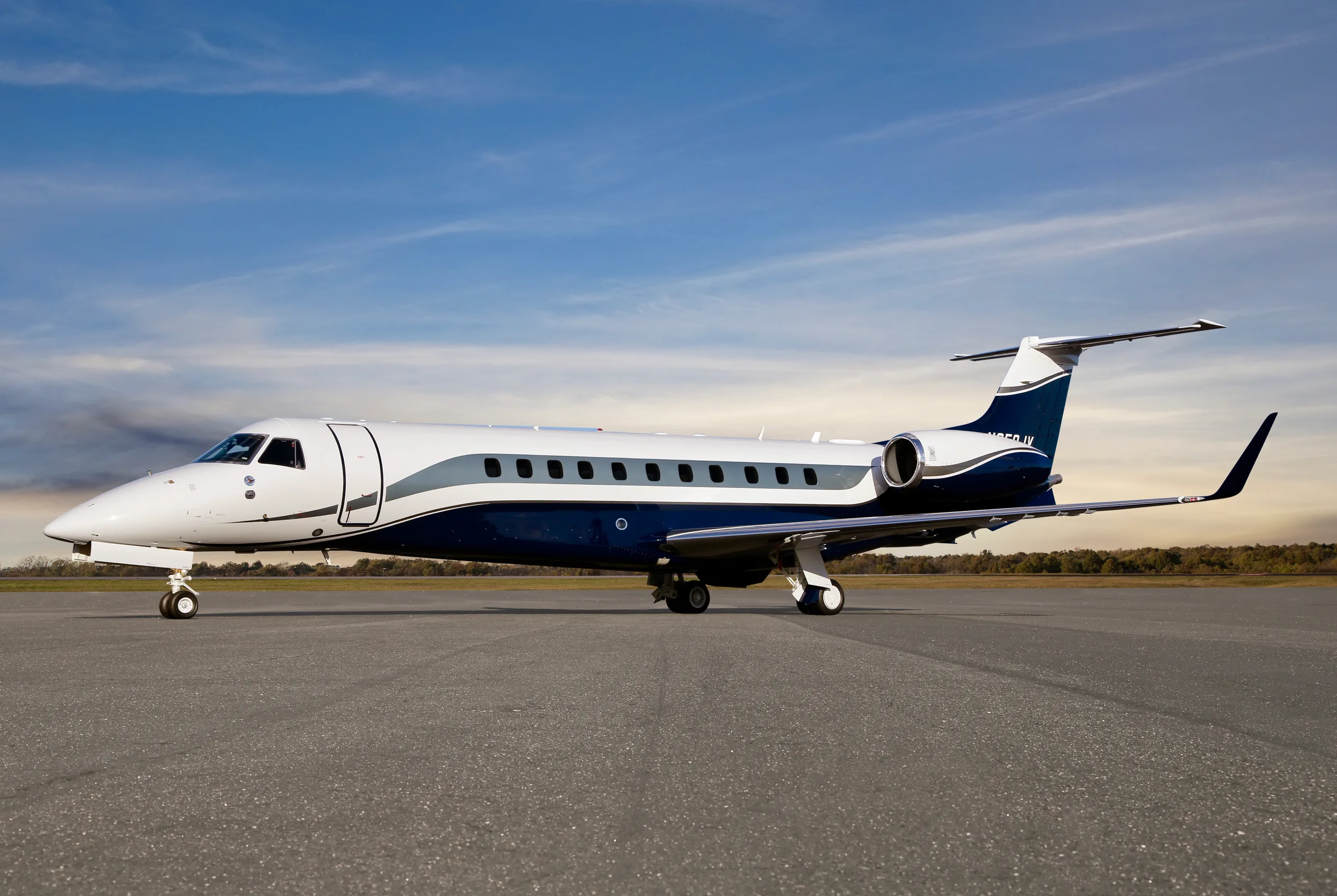 A dark blue, white, and grey Embraer Legacy business jet parked on an airfield under a partly cloudy sky.