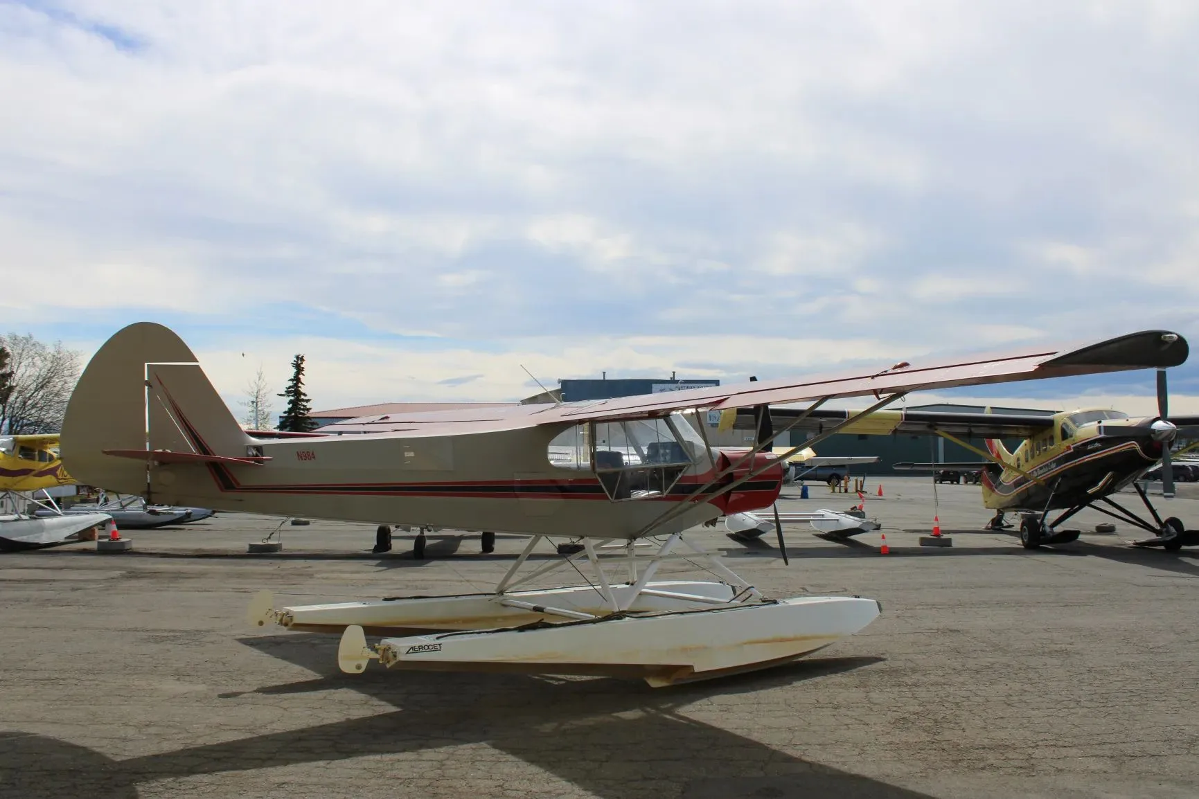 A side view of a red and grey floatplane with N984 on the tail, parked on an apron under a cloudy sky.
