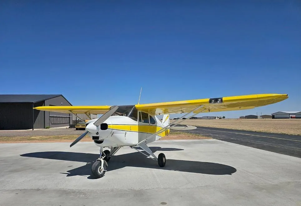 A white and yellow Piper PA-12 Super Cruiser parked on an airfield tarmac with a hangar in the background.