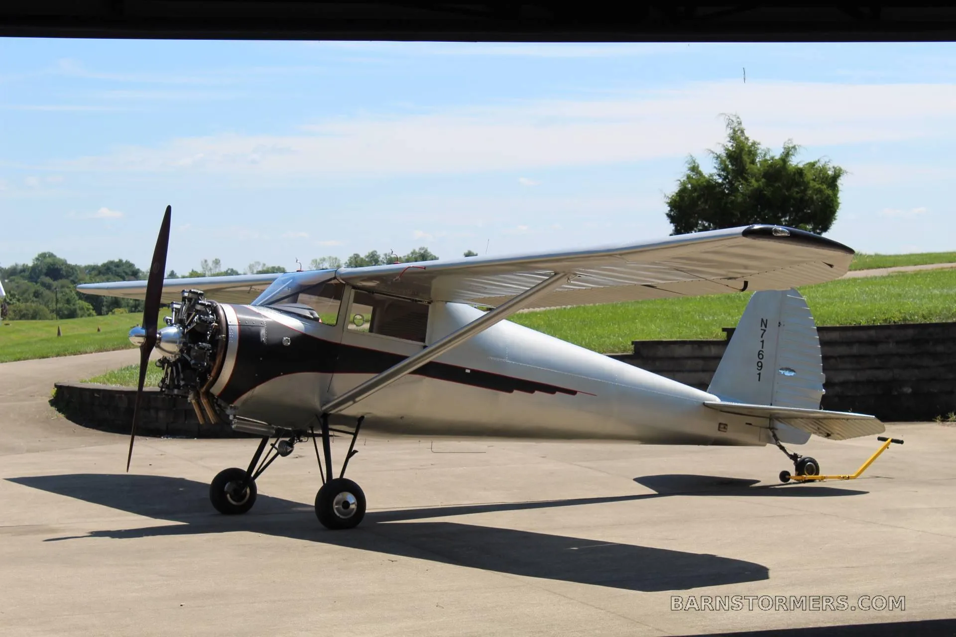 A silver and black high-wing monoplane with an exposed radial engine, tail number N77691, parked on a concrete pad.