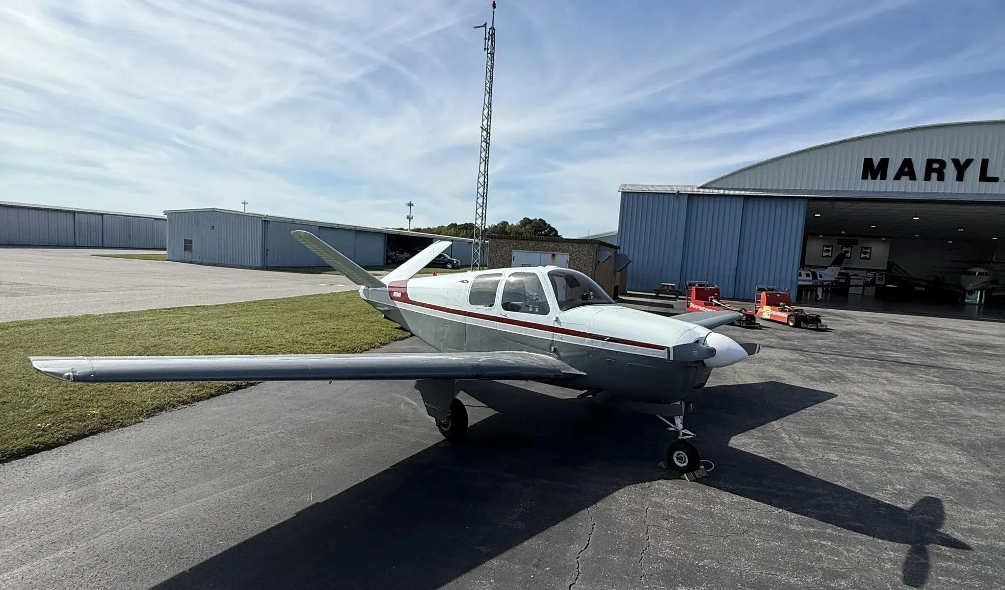 A white and grey Beechcraft Bonanza V-tail aircraft with a red stripe parked on an airfield tarmac in front of a hangar.