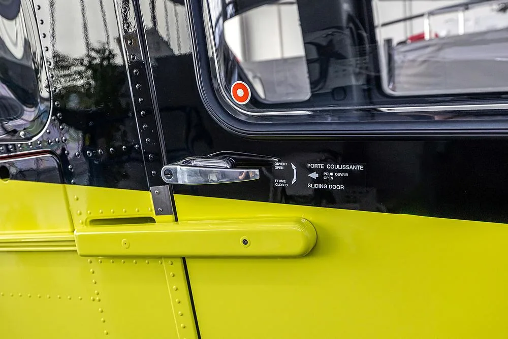 A close-up of the bright yellow and black side of an Airbus H135 helicopter, showing a sliding door handle and instructions.