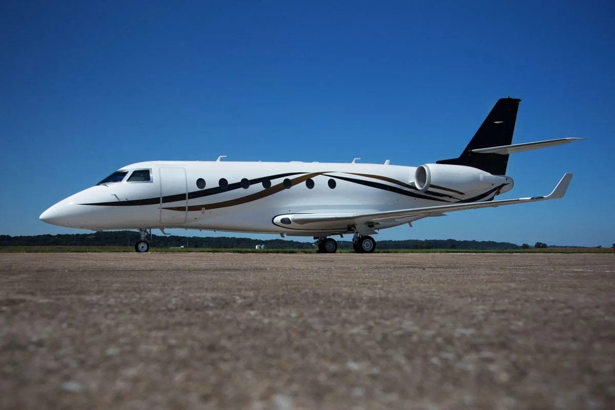 A white business jet with black and gold livery is parked on an asphalt tarmac under a clear blue sky.
