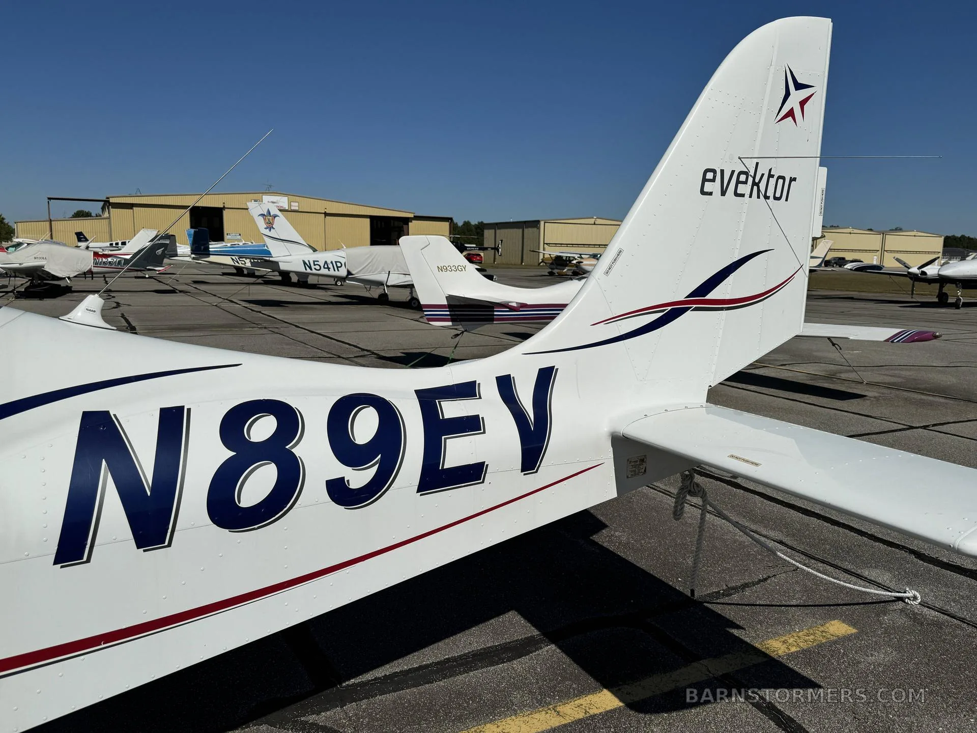 A close-up of the white tail of an Evektor aircraft (N89EV) with blue and red stripes, parked on an airfield.