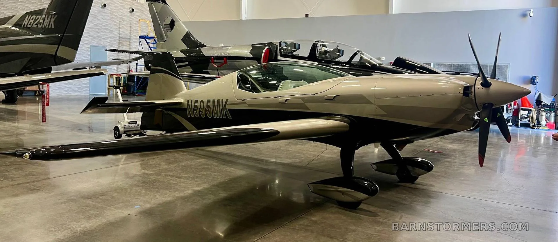 A silver and black propeller aircraft, registration N595MK, parked in a hangar with another jet in the background.