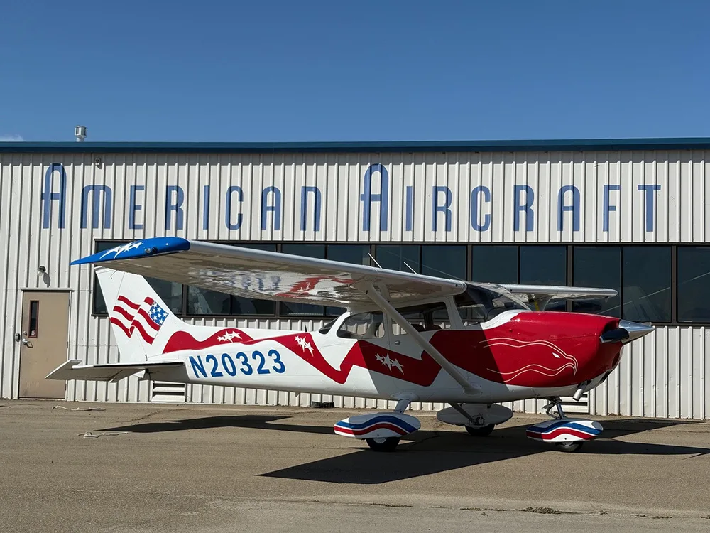 A red, white, and blue Cessna 172 with registration N20323 parked in front of an 'American Aircraft' hangar.