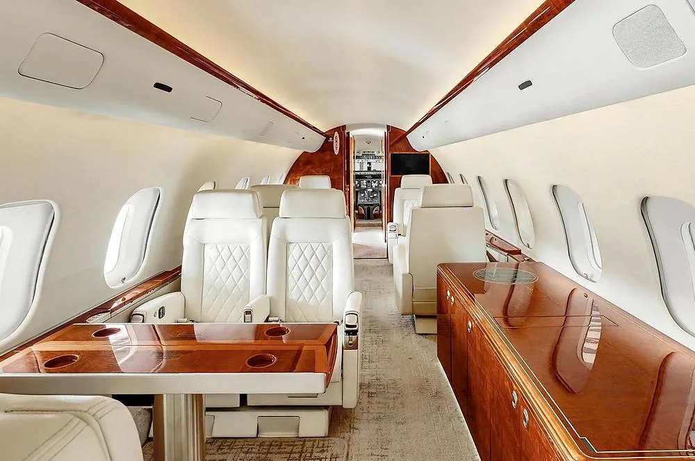 The spacious interior of a private jet cabin with white seats, a large polished wood table, and wood cabinetry.