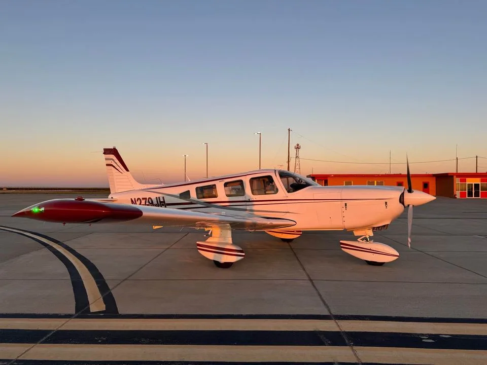A white single-engine propeller aircraft with red accents on the wingtips and tail, parked on an asphalt tarmac at sunset.