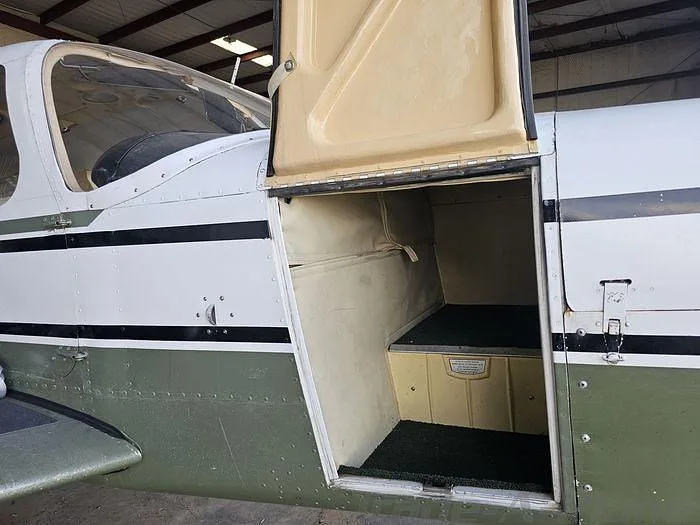 The open baggage compartment of the white and green Piper Cherokee 180, showing its beige interior lining.