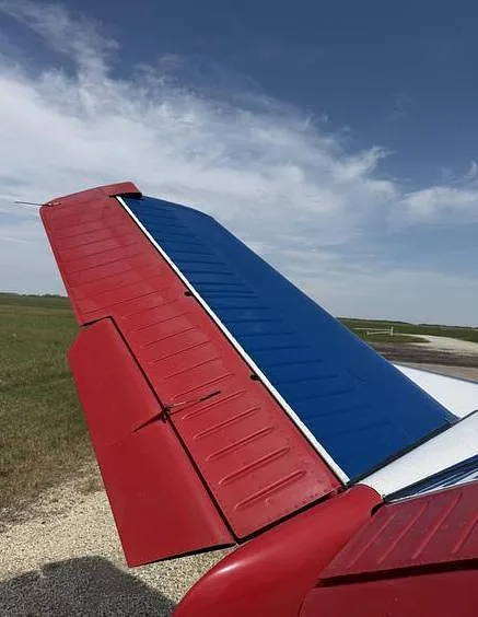 A close-up of the aircraft's red and blue tail section against a blue sky and green field.