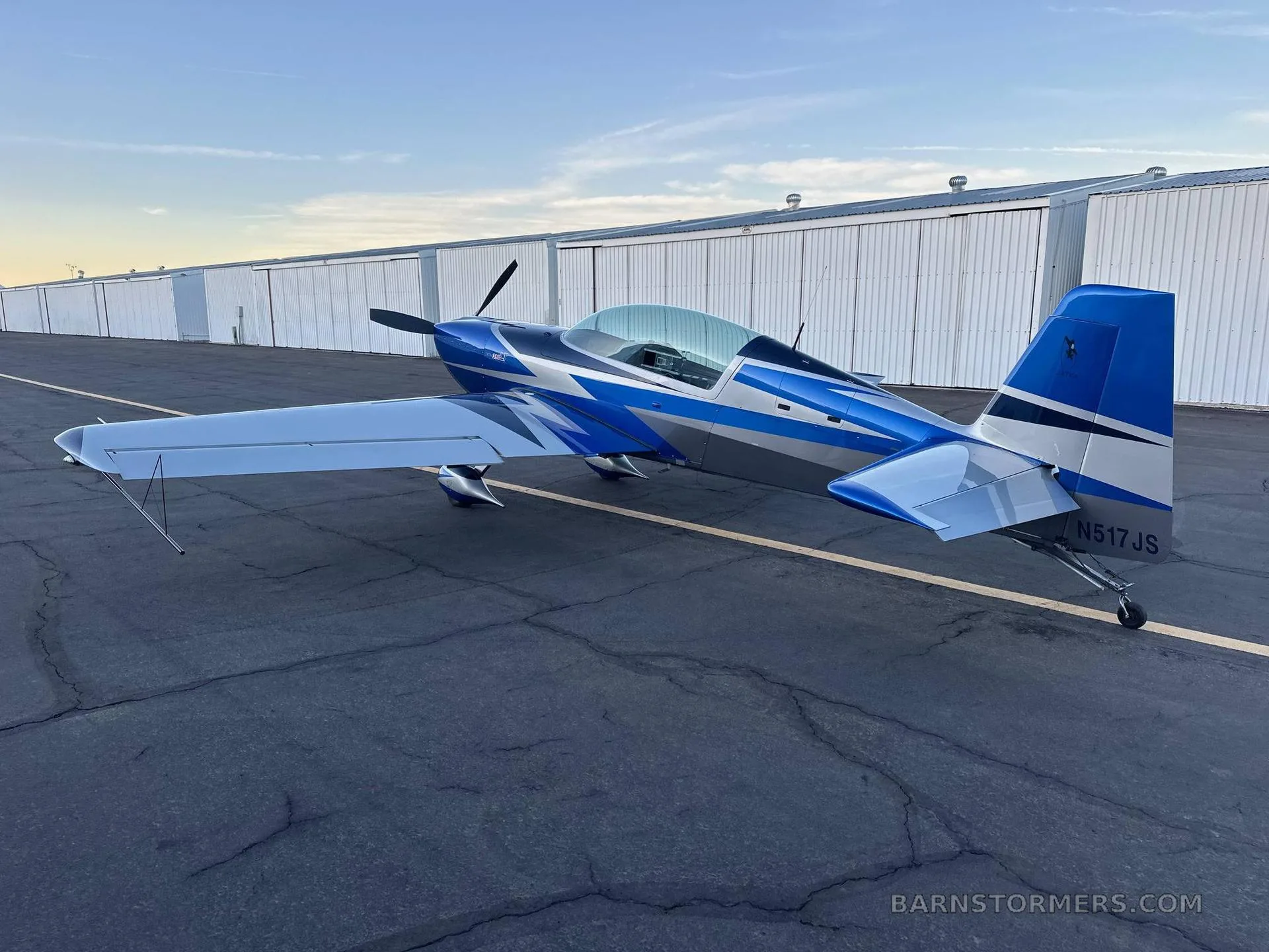 A blue and silver Extra 300LT aerobatic aircraft with tail number N517JS is parked on a tarmac at sunset.