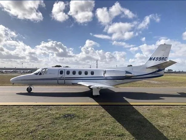 Side view of a white business jet with blue stripes, registration N499RC, on an airfield under a partly cloudy sky.