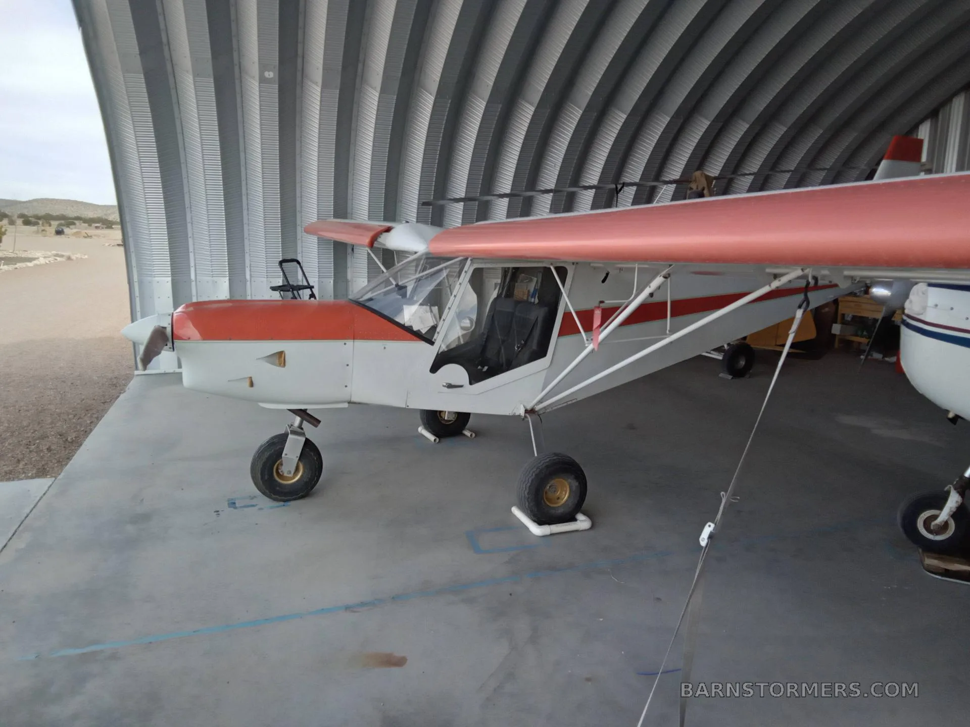 A light sport aircraft with orange and white paint is parked inside a metal hangar, viewed from the side.