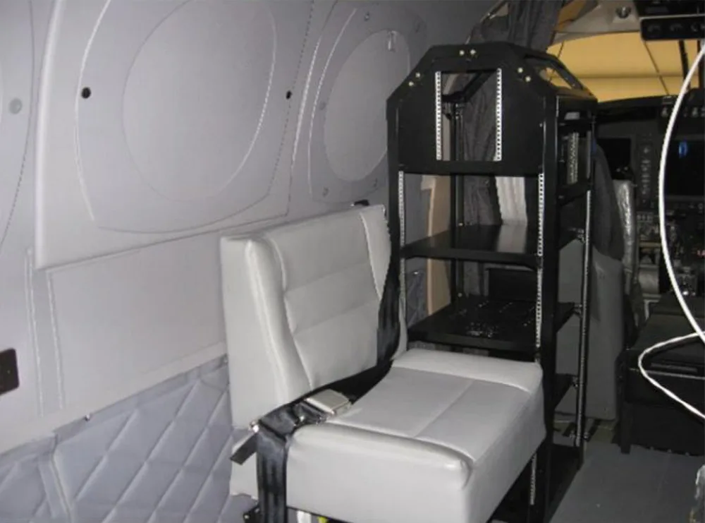 A single grey passenger seat with a seatbelt is visible in the aircraft cabin, next to a black equipment rack.
