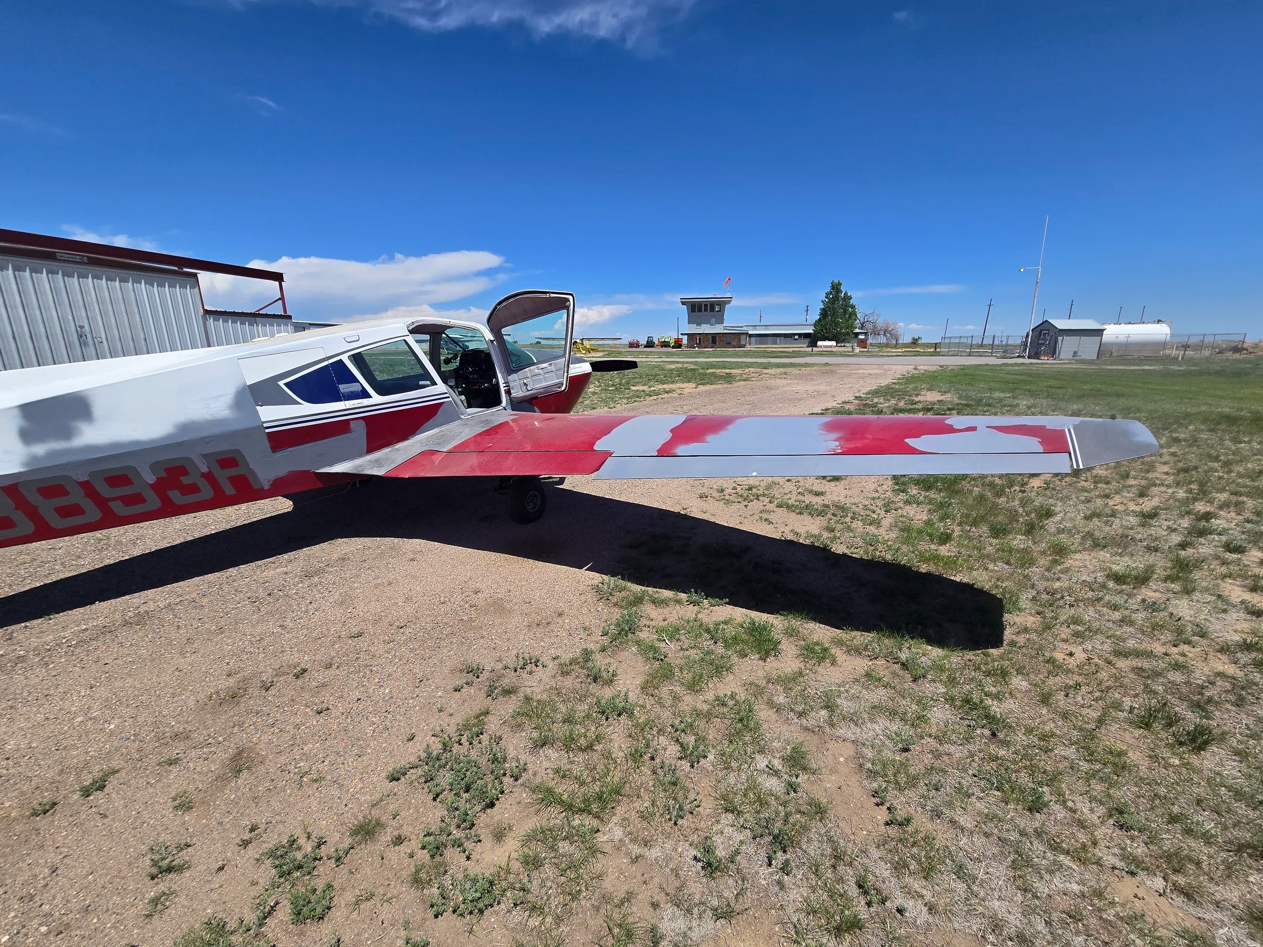 A red and silver single-engine aircraft with its door open, parked on a dirt lot with an airport building in the distance.