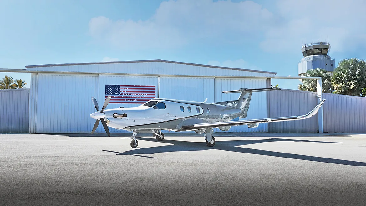 A white and grey Pilatus PC-12 turboprop aircraft, tail number N411PC, parked on an airfield tarmac in front of a hangar with an air traffic control tower in the background.