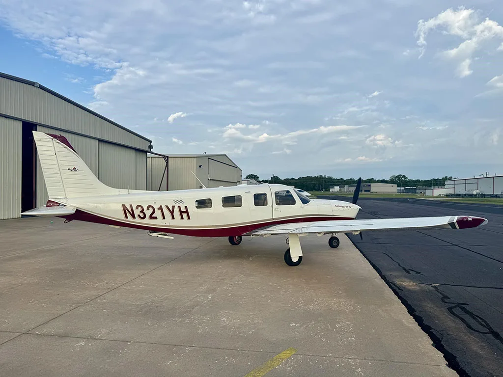 A white and maroon Piper Saratoga II TC aircraft (N321YH) parked on an airport apron next to a hangar under a cloudy sky.