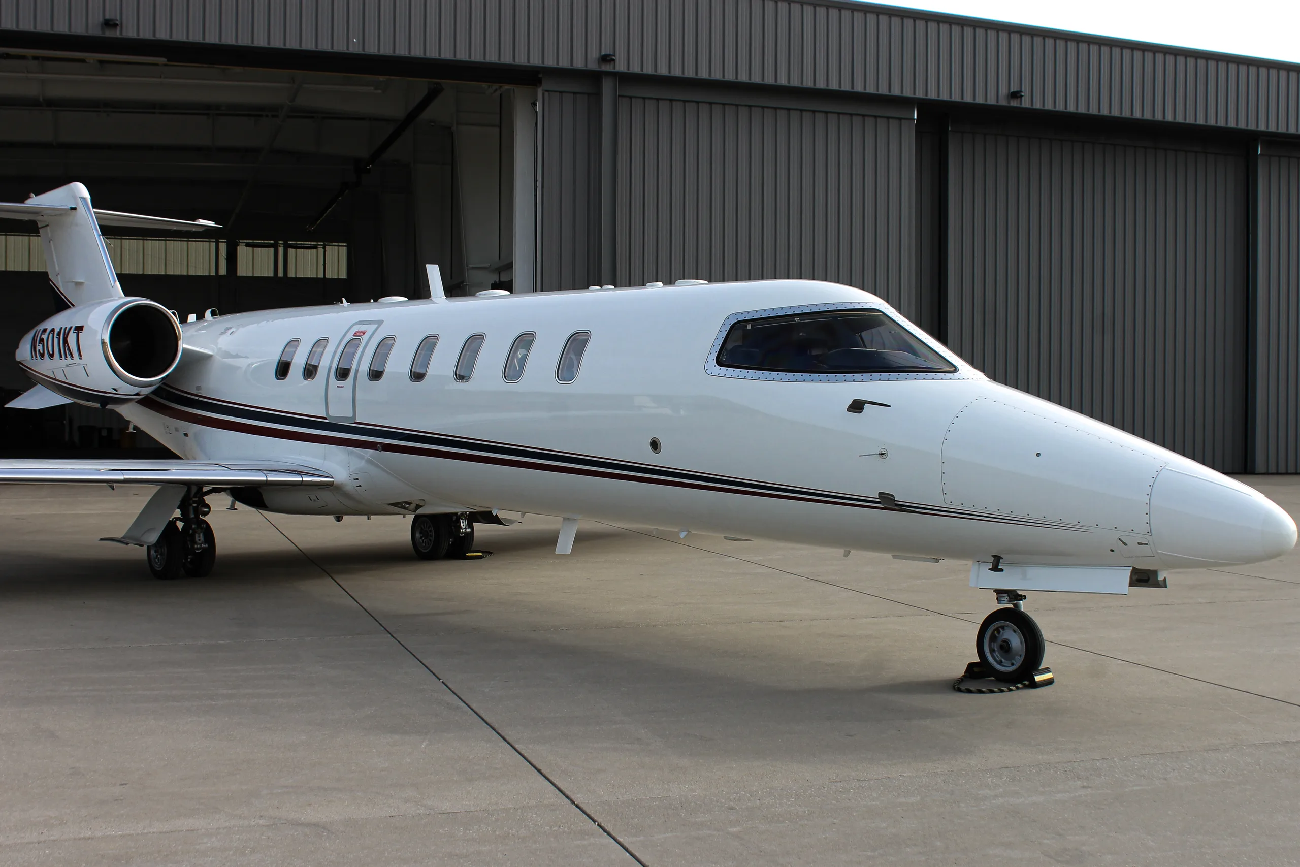 A white Learjet business jet with red and black stripes parked on a tarmac, viewed from the front-right.