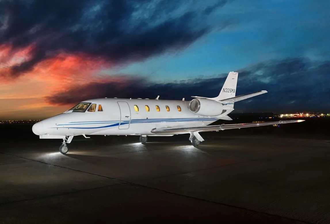 A white Cessna Citation CJ3 with blue stripes is parked on the tarmac at dusk.