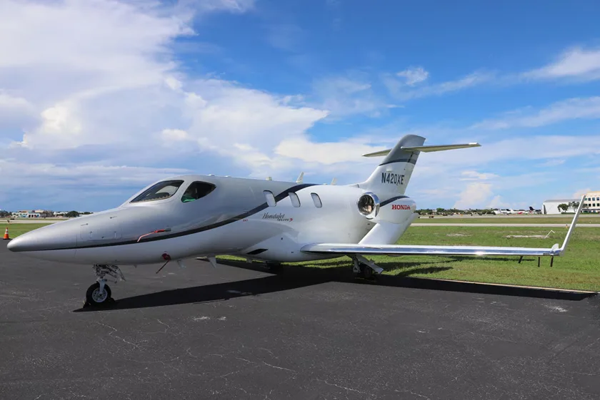 A silver and white HondaJet (N42QXE) is parked on an asphalt apron under a partly cloudy sky.