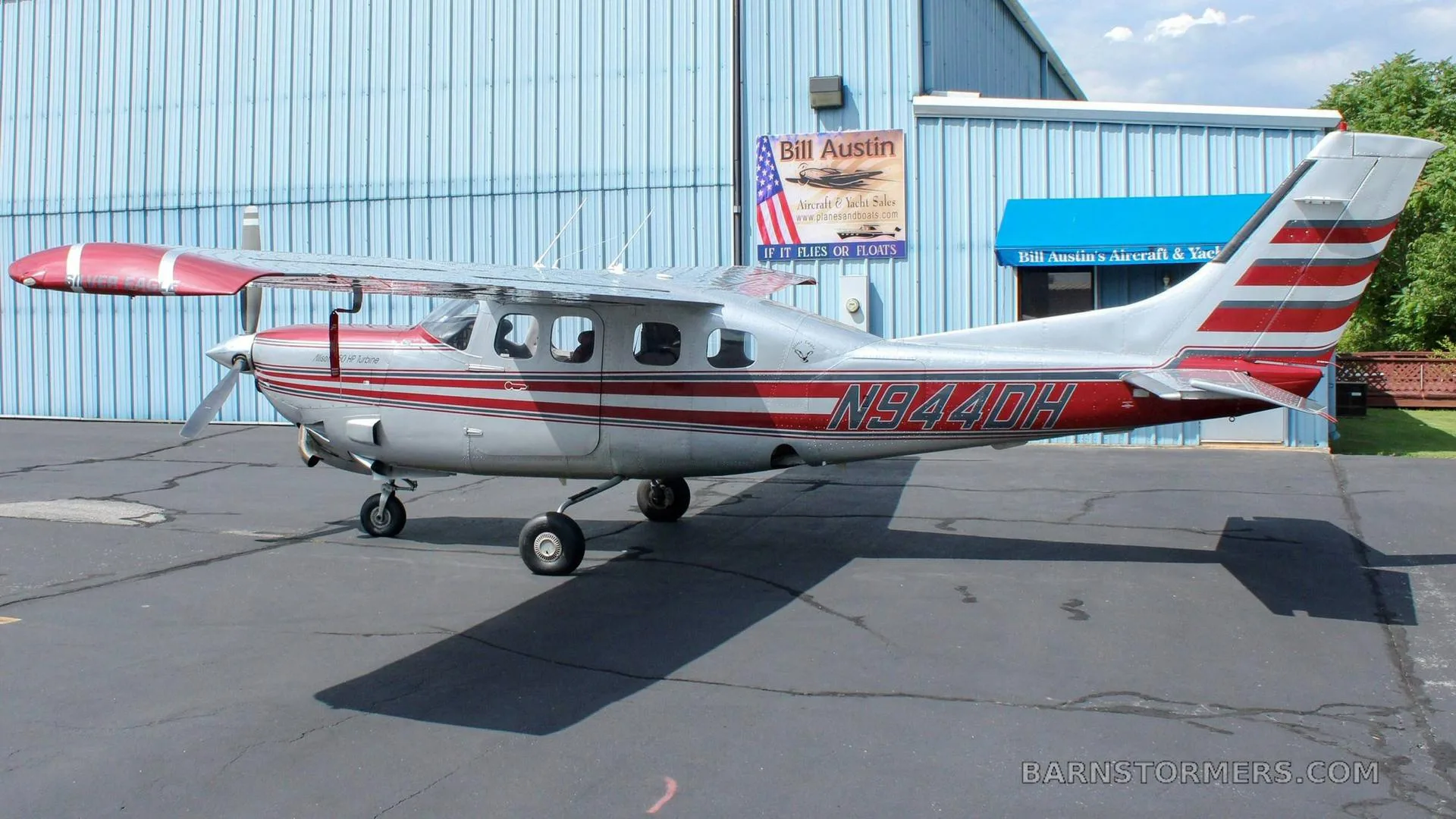 A silver and red single-engine high-wing aircraft with registration N944DH is parked on an asphalt ramp next to a blue hangar.