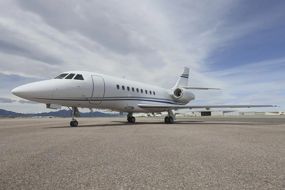 A white Dassault Falcon 900 business jet with blue and grey stripes parked on an asphalt runway, with hangars and mountains in the background.