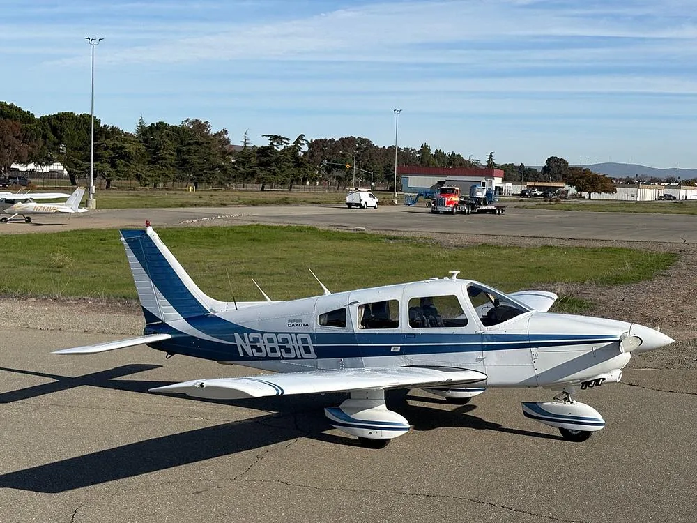 A white Piper Dakota aircraft with blue stripes, registration N3831Q, parked on an airfield with trees and another aircraft in the distance.