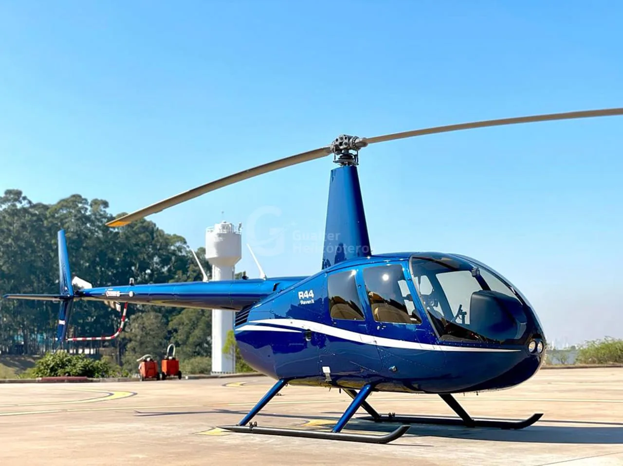 A blue Robinson R44 Raven II helicopter with white stripes parked on a helipad under a clear sky.