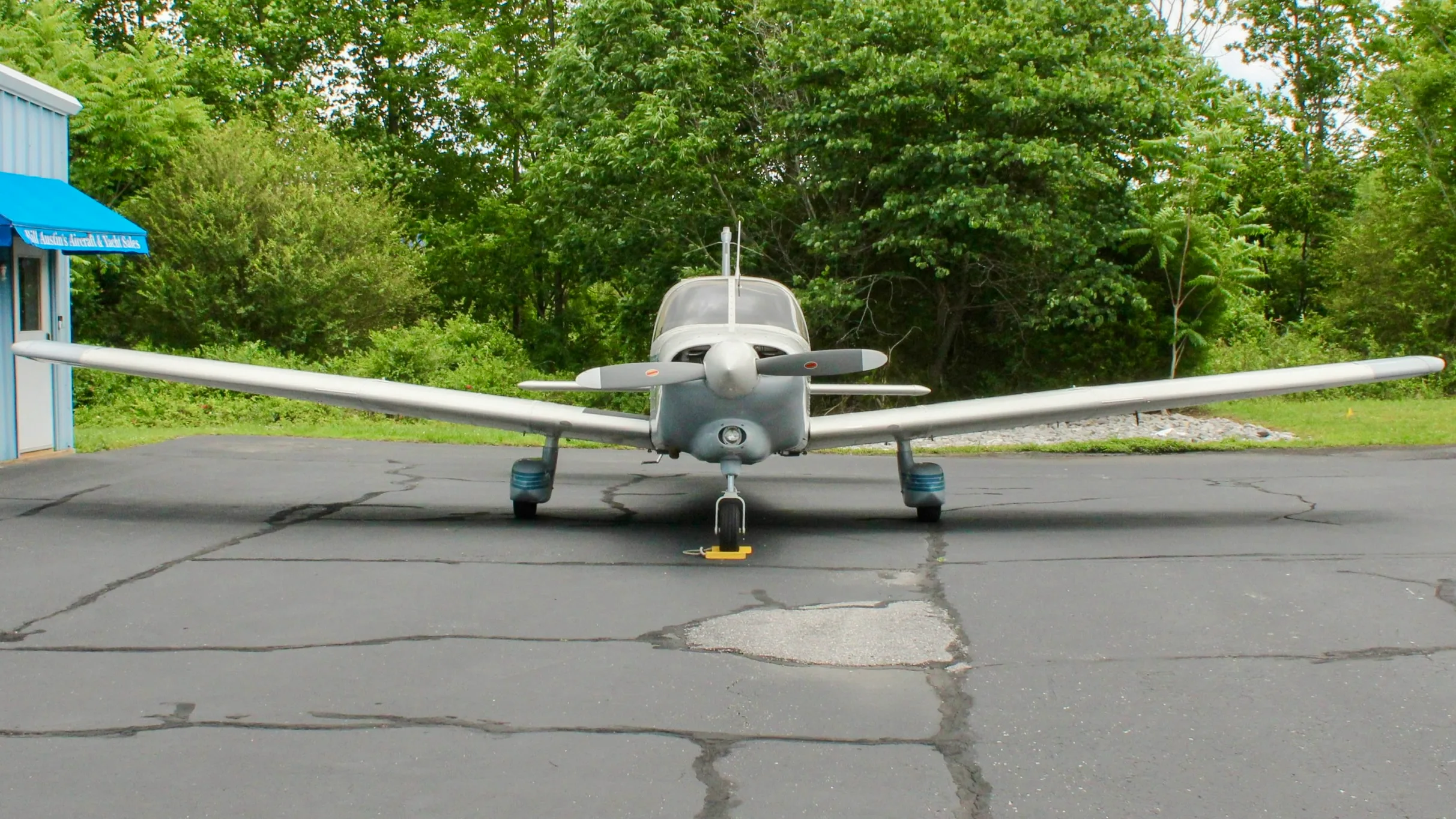 A front view of a white and blue Piper Cherokee aircraft with a single propeller, parked on an asphalt ramp.