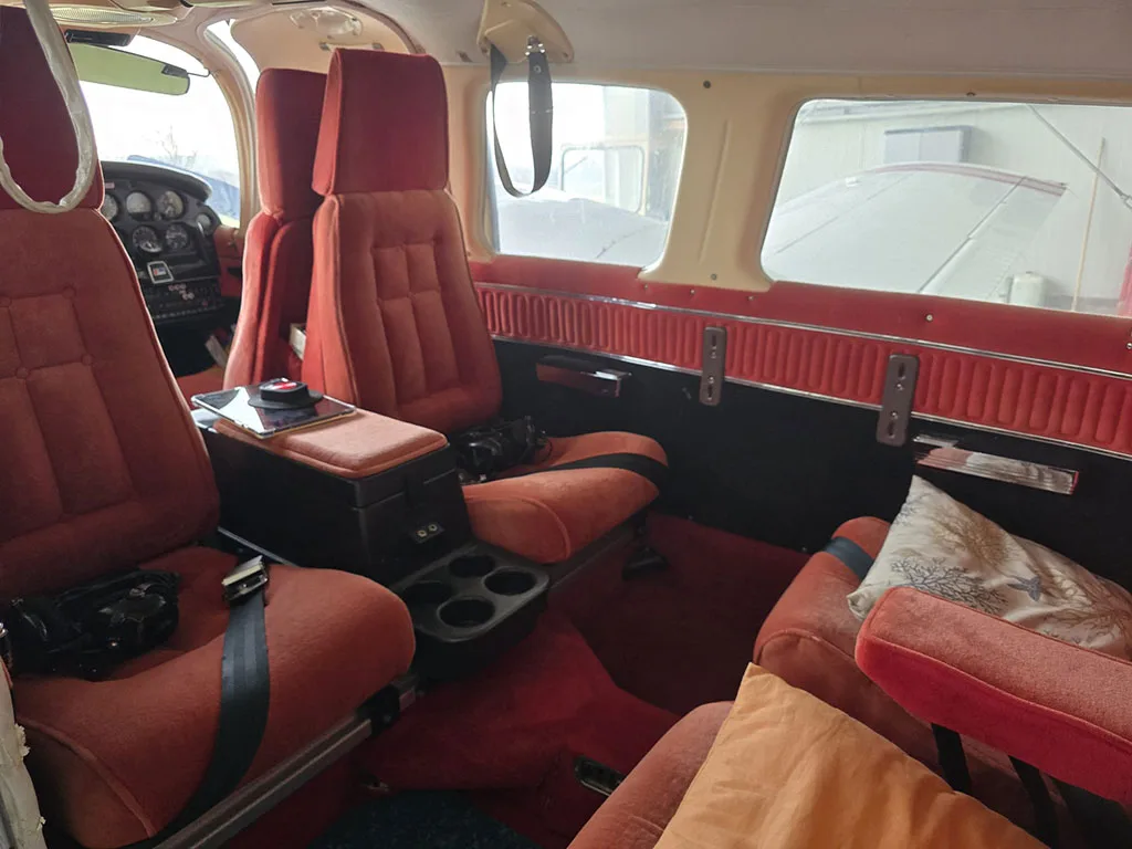 The aircraft's interior features red upholstered seats, a central console with cup holders, and a view of the wing outside the window.