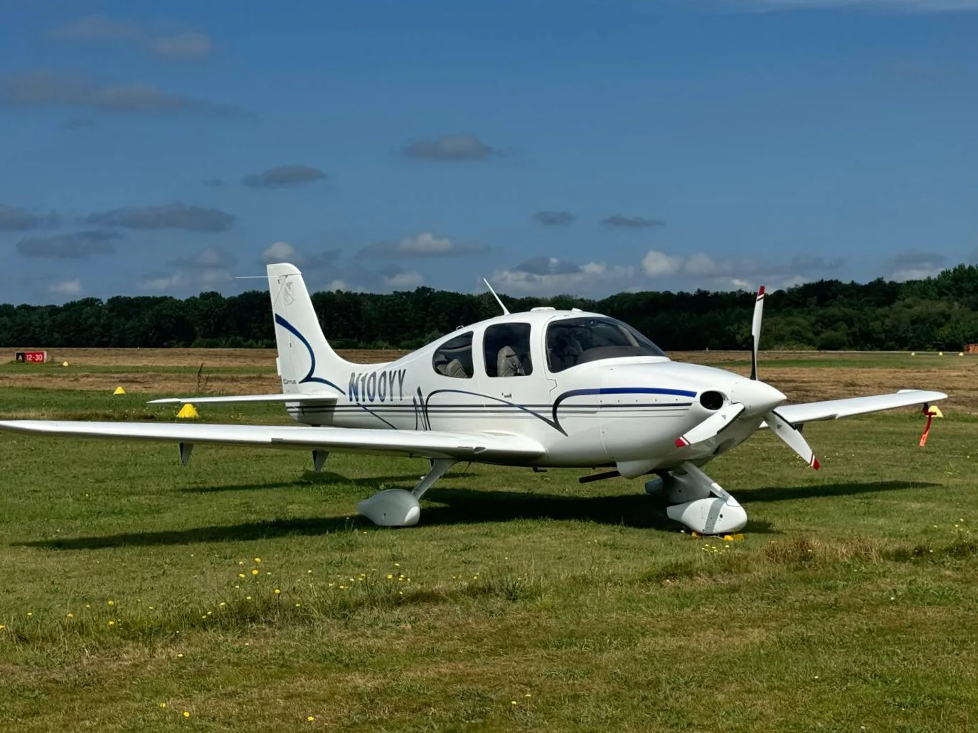 A white Cirrus SR22 aircraft with blue and grey stripes parked on a grassy airfield under a blue sky.