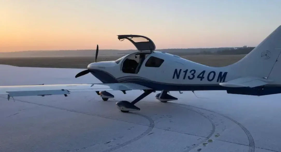 A white and blue Columbia 400 aircraft with registration N1340M parked on a snowy field at sunset, with its door open.