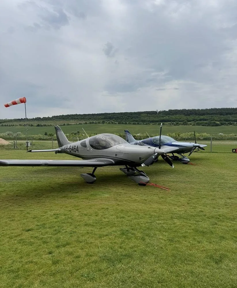 Two light aircraft, one silver and dark grey (YR-6464), parked on a grassy field with a windsock.