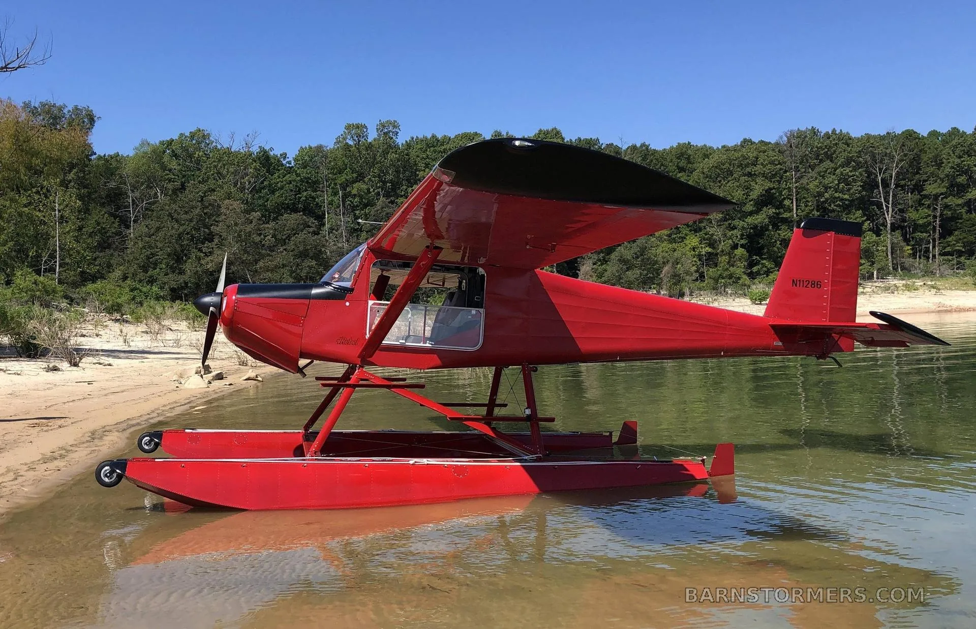 A red floatplane with registration N11286 is partially in the water and on a sandy beach next to a forest.