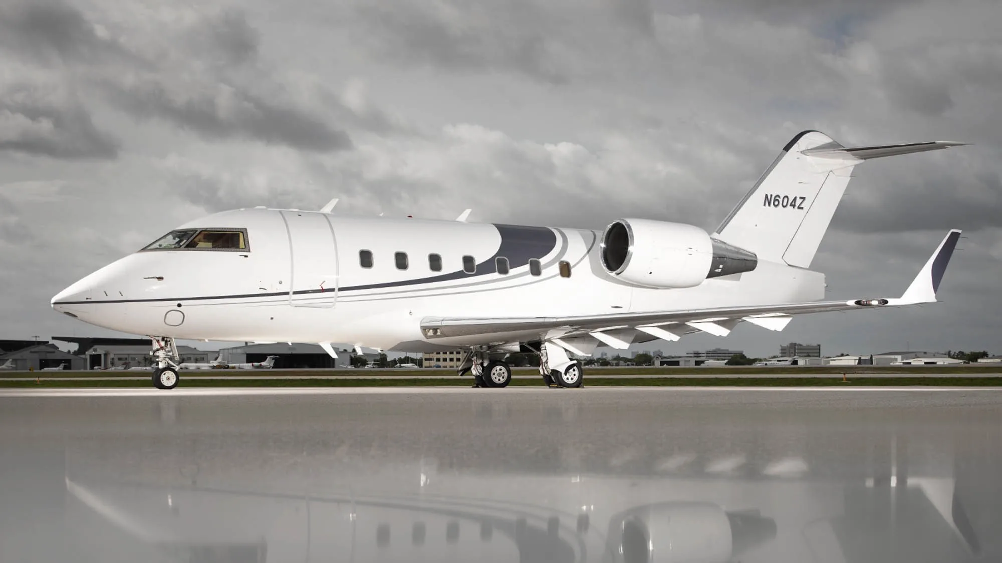 A white Bombardier Challenger 604 business jet with dark grey stripes, registration N604Z, parked on a wet tarmac under a cloudy sky.