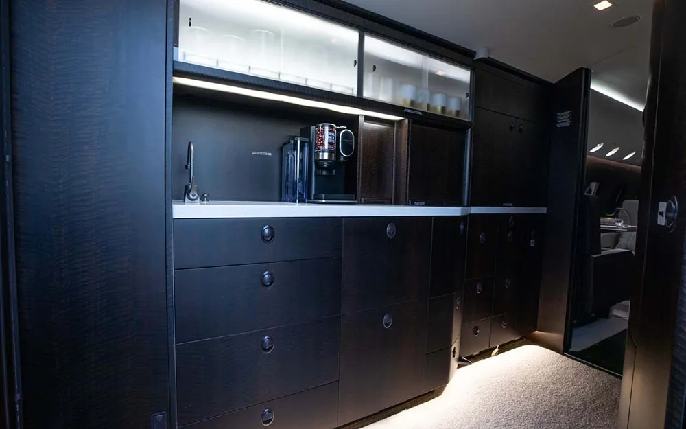 The dark wood galley area of a private jet, equipped with a coffee machine, sink, and storage cabinets.
