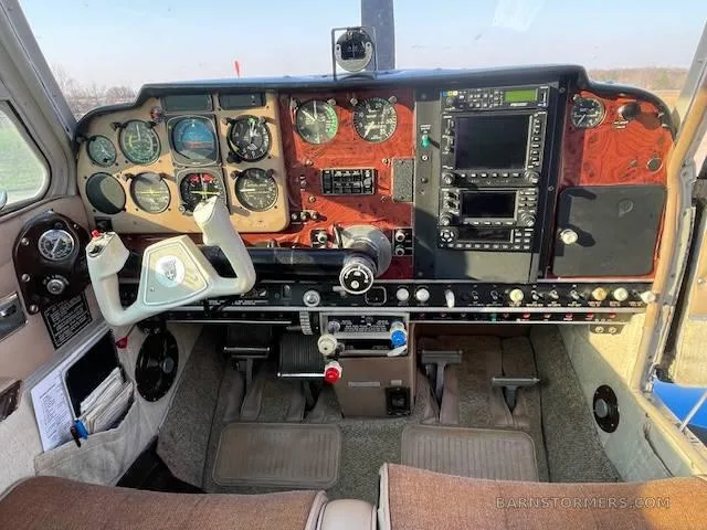 The cockpit of a small aircraft features a wood-grain instrument panel with various gauges, screens, and a white control yoke.